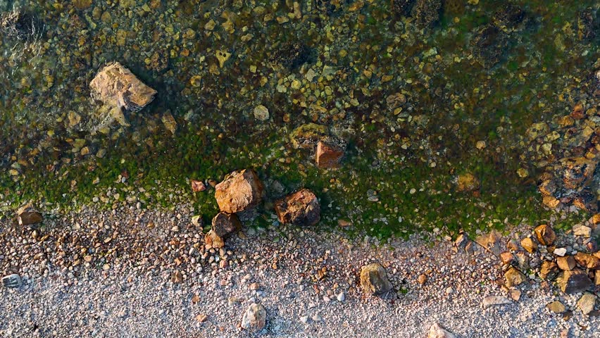 An aerial shot looking down at a calm shoreline where clear water gently laps at a beach made of smooth pebbles and larger rocks a serene and peaceful coastal scene video