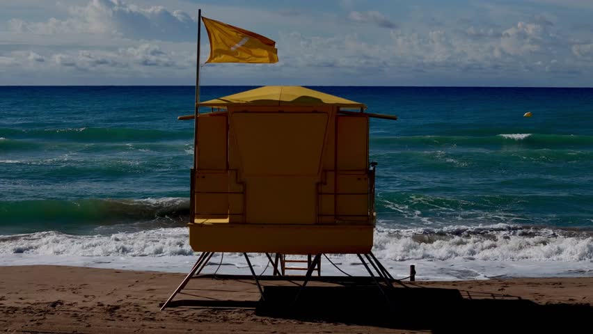 A drone shot of a yellow lifeguard tower with safety flag waving in the wind, facing the turquoise Mediterranean Sea under a cloudy sky in Spain