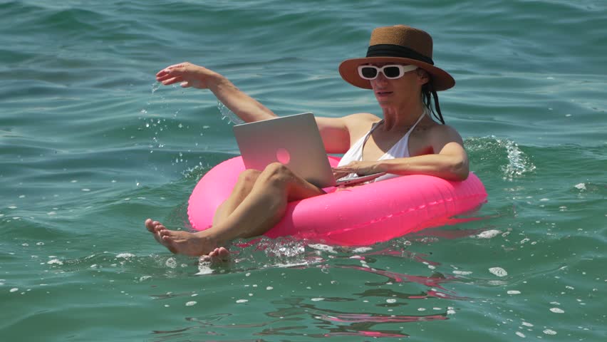 Woman Laptop Water: Woman works on laptop floating on pink inflatable in the ocean for remote work, bright sunny day.
