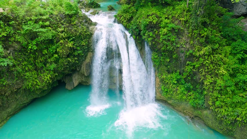 Aerial view of a powerful waterfall cascading into a vibrant turquoise pool, surrounded by lush green tropical foliage, with a person swimming, Tavoro Falls, Fiji.