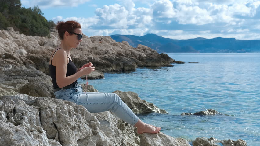Woman knitting on rocks by the sea enjoying sunny day. Young woman relaxing near ocean, sitting on rocky coastline, knitting with vibrant red yarn under bright sunlight, wearing sunglasses