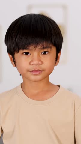 Cheerful Asian boy in beige shirt points and smiles playfully at camera indoors, blurred background.