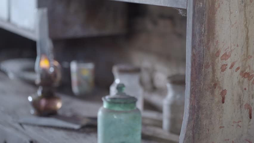 A small oil lamp burning on a dirty table among an old ice saw, jars, and glasses, with soft shifting focus and artistic lighting.