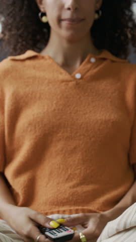 Vertical cropped shot of young woman sitting comfortably on sofa at home, turning on TV and flipping channels with remote during leisure time