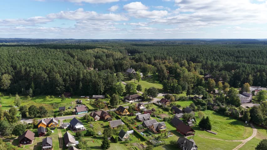 Drone view of Palūšė village in Lithuania with traditional houses, fields, and a historic wooden church surrounded by dense green forest under a partly cloudy sky.