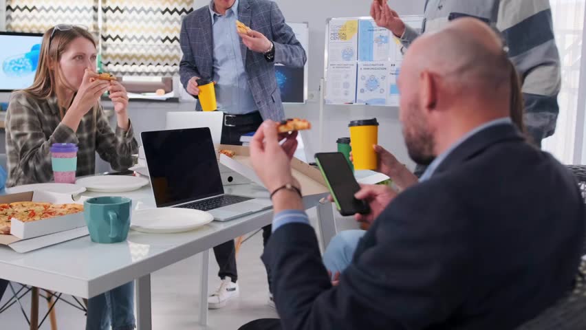 Group of colleagues in casual and business clothes taking a lunch break at the office table, eating pizza and drinking coffee. Informal teamwork atmosphere, laptops and documents visible.