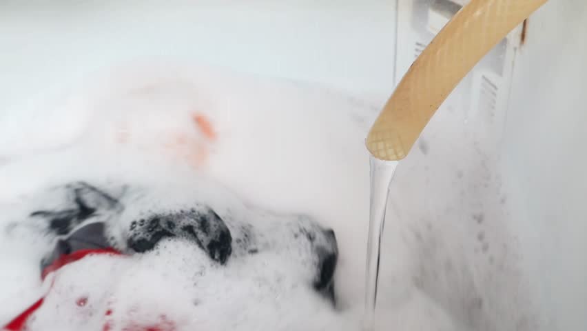 Water flows from a hose into a washing machine filled with colorful clothes and soap suds, laundry and household cleaning process.