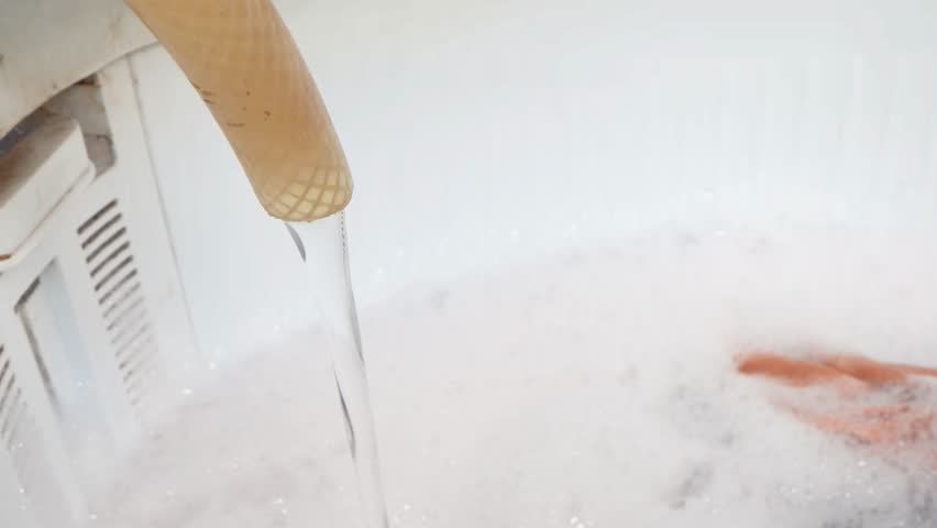 Water flows from a hose into a washing machine filled with colorful clothes and soap suds, laundry and household cleaning process.