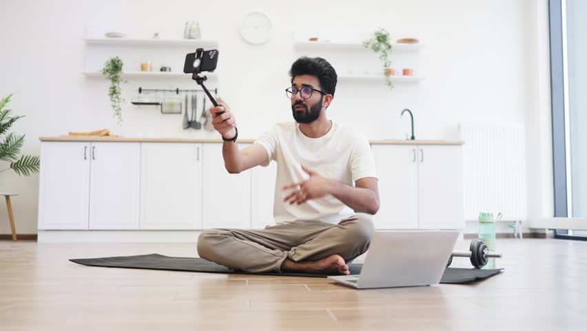Young adult man recording video blog about fitness and health at home, sitting cross-legged on mat, using smartphone mounted on stick, with laptop and water bottle nearby in bright kitchen setting.