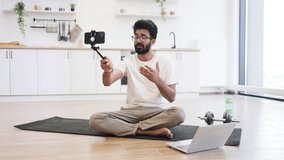 Young adult man sitting on yoga mat creating fitness and health video blog using laptop and phone in modern home environment. Promoting wellness and exercise culture. Bright and clean indoor setting. - Powered by Shutterstock - Get 15% off with code: PIKWIZARD15