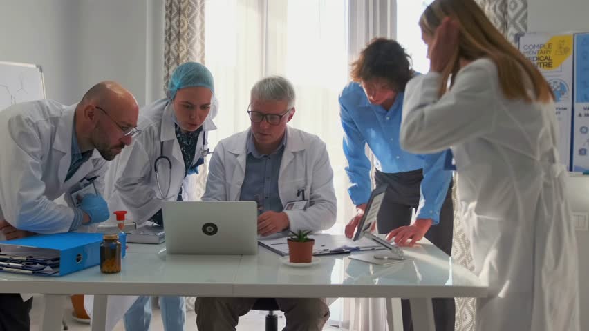 Group of doctors in lab coats actively debating medical research and scientific reports. Senior physician leads discussion on suitability of new drugs. Team studies data and clinical papers in office.