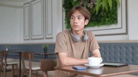 Young man in cafe restaurant using digital tablet computer while thinking he is wearing casual t-shirt and headphones around his neck and has coffee cup on table with mobile phone - Powered by Shutterstock - Get 15% off with code: PIKWIZARD15