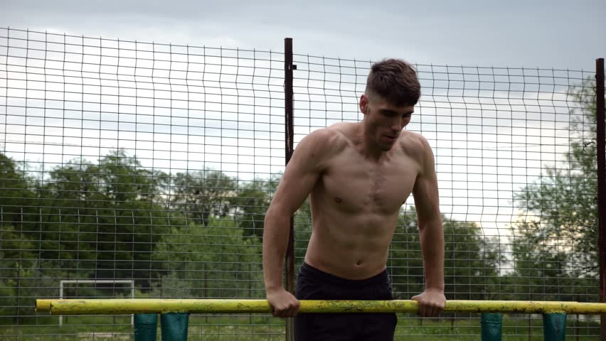 Male shirtless athlete doing push ups on parallel bars at nature. Strong sporty man exercising at sports ground. Young sportsman training hard outdoor. Concept of active sportive lifestyle. Slow mo