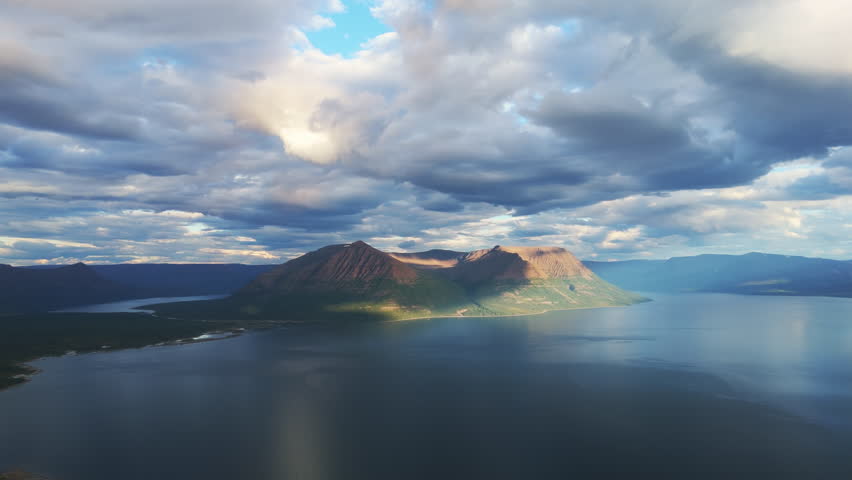 Sunlit island mountain rises from Lake Lama under dramatic cloud, Putorana Plateau, Russia. Evening sun draws warm highlight and cool shadow across the ridge and calm water, ideal for travel promotion