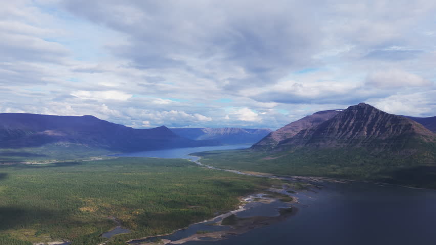 Cloud shadow sweeps across the bay of Lake Lama while a stratified peak of the Putorana Plateau dominates the frame. Shoreline, taiga forest and quiet water define a remote destination