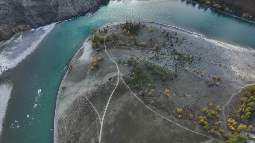A high-angle aerial view of a large, teardrop-shaped gravel island at the confluence of two rivers, with dirt tracks and a few autumn trees