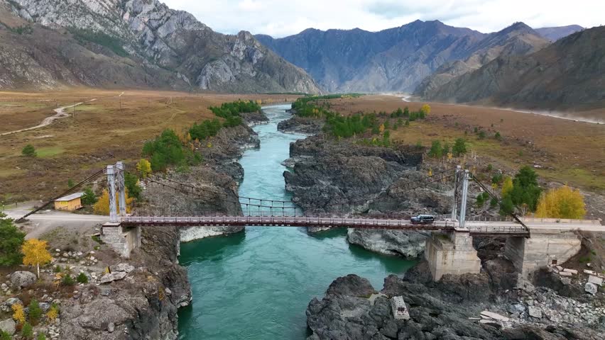 An aerial view of a car crossing a metal suspension bridge over a vibrant turquoise river, with rocky shores and a wide valley surrounded by mountains in the distance