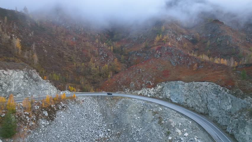 An aerial view of a car driving on a winding paved road that hugs the side of a steep mountain, with rocky slopes and a foggy sky, showing the challenging and adventurous nature of the journey