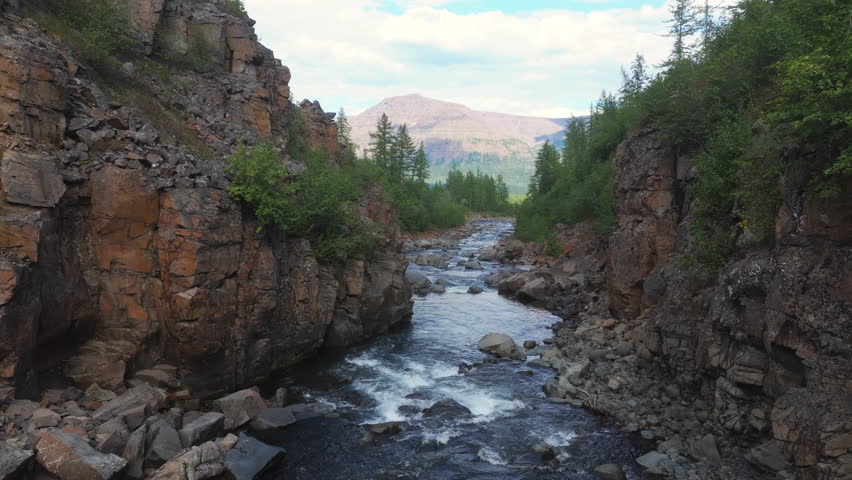 Steep basalt wall frames a narrow river as the drone glides upstream through a rocky canyon near Lake Lama. Summer light reveals clear water and distant mountain, inviting travel and adventure