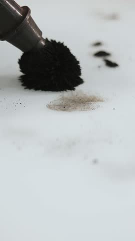 Forensic brush revealing a clear fingerprint with black powder during crime scene investigation. Vertical close up of fingerprint development on surface.