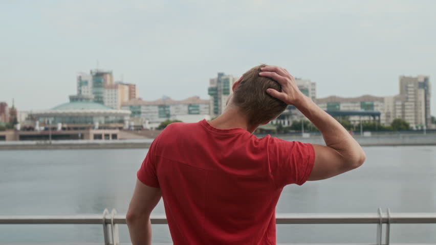 Back view of unrecognizable athletic man warming up neck and back muscles while standing on promenade with buildings and river in background