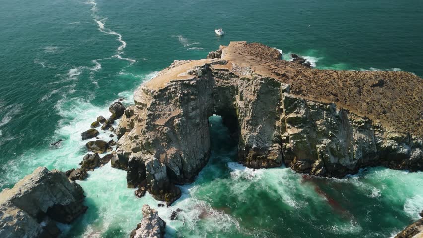 Aerial shot rises and orbits around massive natural sea arch at Palomino Island, flock of birds circling and tour boat moored nearby against turquoise ocean