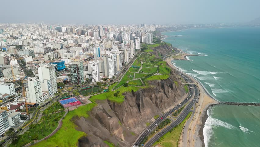Famous Costa Verde in Lima, Peru, showcasing the dramatic cliffs, modern cityscape of Miraflores, and busy coastal highway next to the Pacific Ocean - aerial flyover
