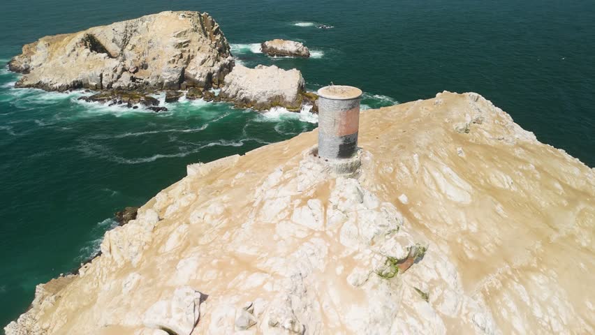 An aerial parallax shot orbits an old stone lighthouse on the summit of the Palomino Islands in Peru, with numerous seabirds flying through the dramatic coastal scene