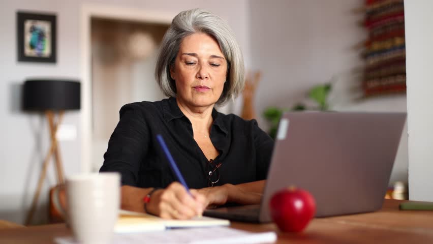 Professional senior businesswoman writing notes while working remotely on laptop at home office, demonstrating focused concentration and calm productivity