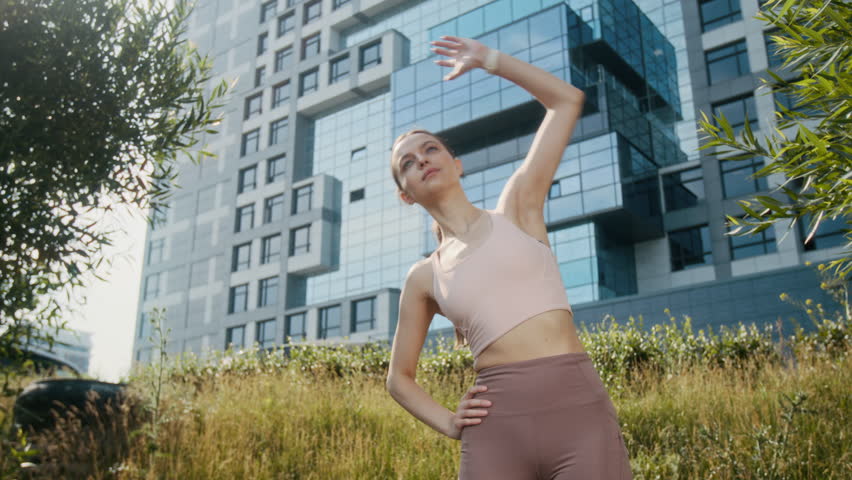 Low angle of young brunette Caucasian woman doing side bends while doing stretch exercise in park, modern building raising in background