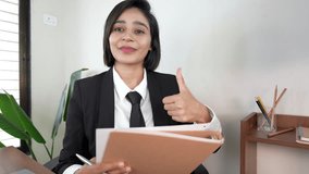 A confident young Sough Asian businesswoman in suit smiling, holding file, giving thumbs-up at office desk - Powered by Shutterstock - Get 15% off with code: PIKWIZARD15