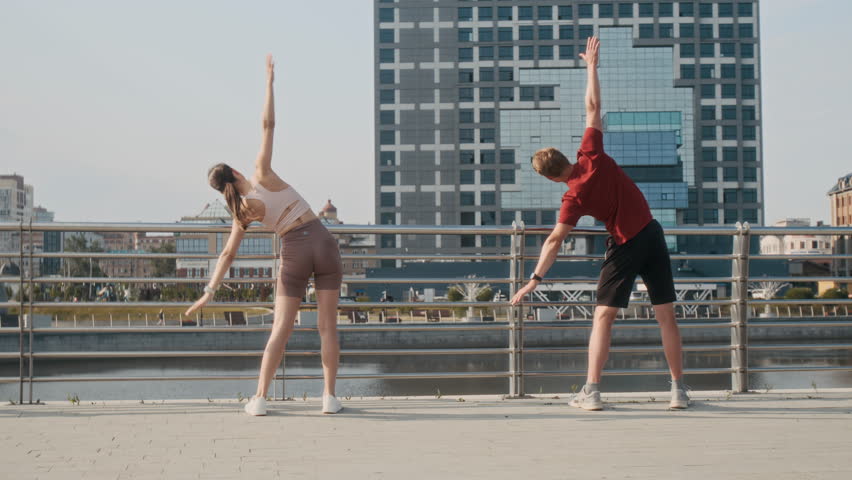 Back view of brunette woman and blond man doing side bends while preparing for jogging time on promenade
