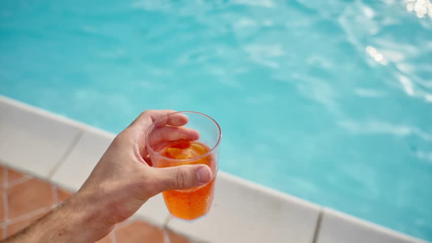 Point of view of hand holding fresh orange drink by swimming pool and drinking. POV man enjoying cocktail in pool party in backyard home or hotel resort during summer vacation holiday hot sunny day