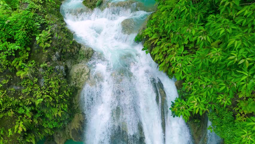 Aerial view of Kawasan Falls, a multi-tiered waterfall cascading into a turquoise pool, surrounded by lush tropical jungle with people swimming, in Cebu, Philippines.