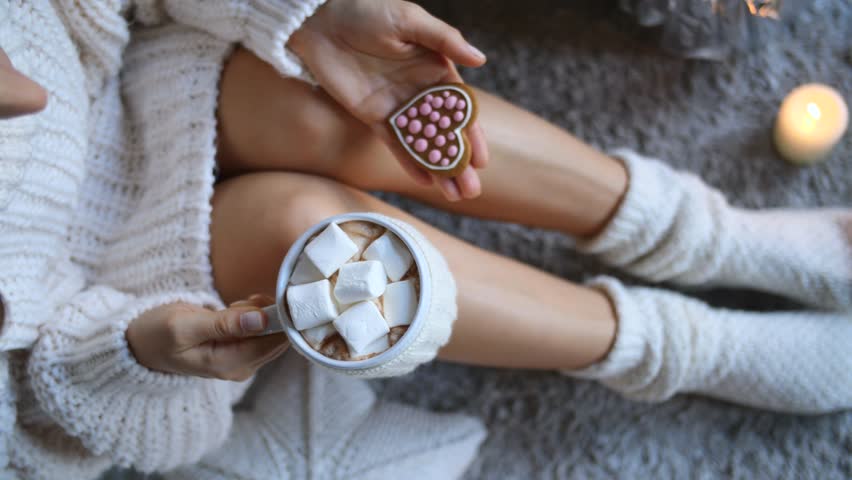 Cozy winter scene featuring a woman wearing a white sweater and socks, relaxing with a mug of hot chocolate with marshmallows and a heart shaped cookie, a lit candle providing a warm ambiance