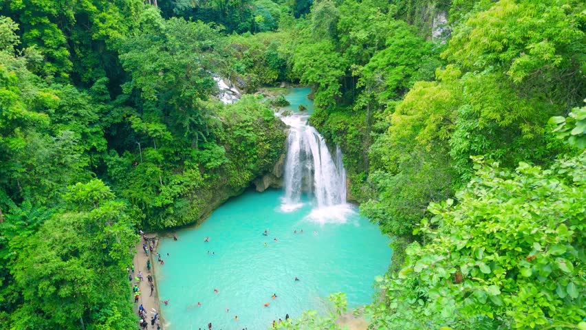Aerial view of Kawasan Falls, a multi-tiered cascading waterfall with clear turquoise pools. Individuals swim and enjoy the lush tropical jungle surroundings in Badian, Cebu, Philippines.