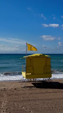 A vertical view of a solitary yellow lifeguard hut with a waving flag, placed centrally on a wide beach with turquoise sea and blue sky