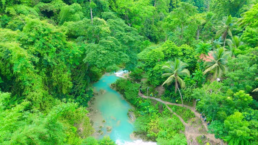 Aerial view of a turquoise river flowing through a dense tropical jungle, with small waterfalls and people hiking along a path at Kawasan Falls, Philippines.