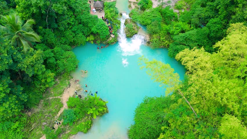 Aerial view of a turquoise waterfall cascading into a pool, surrounded by green jungle. Several men, women, and young adults swim at Kawasan Falls, Philippines.