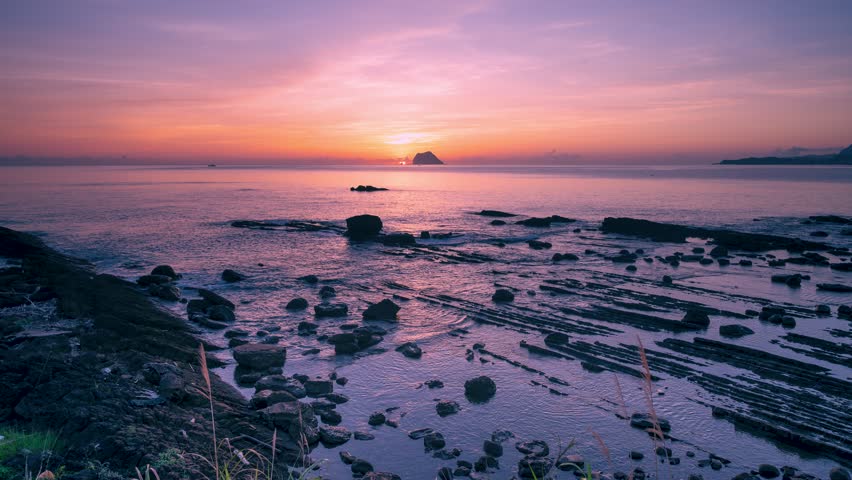 Early autumn dawn at Wanli, Taiwan. The sun rises behind Keelung Islet, casting a dreamlike glow over the sea with pink and orange skies and rocky shores.