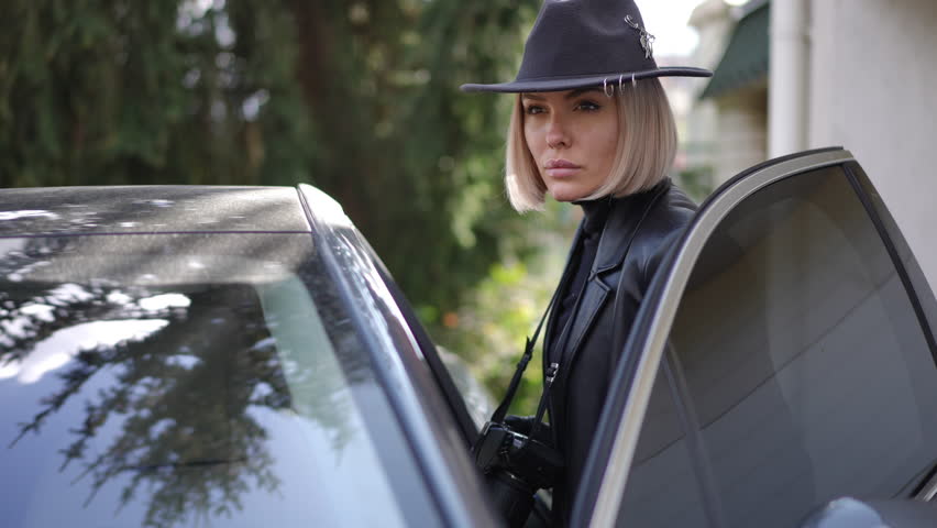 A female detective in black leather coat and hat secretly observes surroundings with camera from car, documenting evidence for investigation.