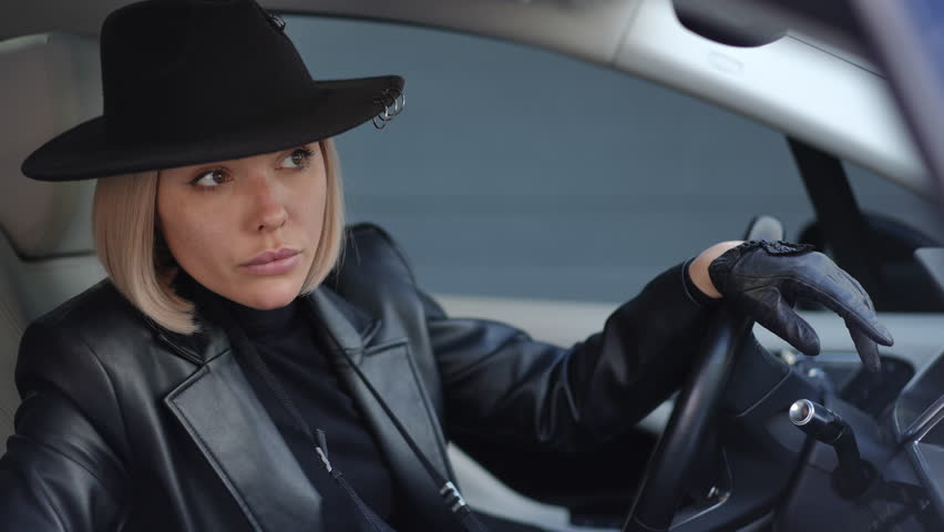 Female detective in leather jacket and black hat holds camera inside car, watching closely during investigation work.