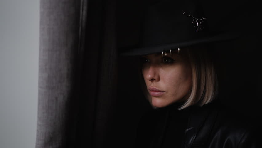 Female detective in black hat and leather outfit secretly watches through a curtain in a dimly lit room, observing the scene with focus.