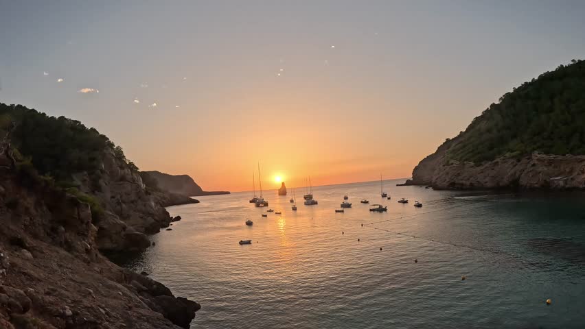 Scenic sunset over bay in Ibiza, Spain, with yachts and boats anchored on calm Mediterranean Sea surrounded by rocky cliffs, creating peaceful seascape and travel atmosphere in summer evening.
