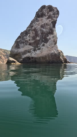 Rock ocean reflection, large formation in calm water under clear sky, beautiful seascape, vertical video.