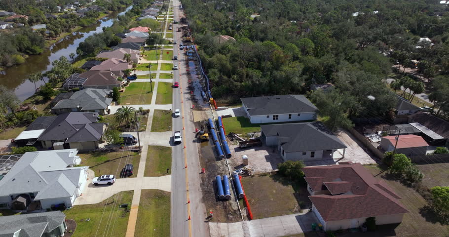 Florida neighborhood street under repair, with one lane closed by cones and temporary lights while vehicles navigate past roadwork equipment.
