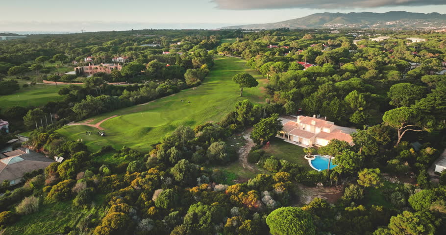 Portugal, Lisbon: Aerial view golf course in Cascais with lush green meadow ad trees landscape, luxury villas houses, mountain and ocean in background. Beautiful sunset scene. Drone flight footage