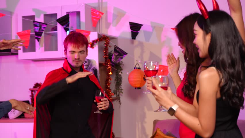 Asian friends in creative Halloween costumes gather on a sofa under colorful RGB lights, laughing and enjoying a festive night party filled with spooky decorations, joy, and playful holiday spirit.