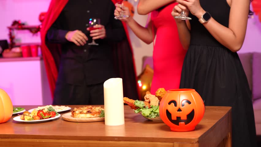 Asian friends in creative Halloween costumes gather on a sofa under colorful RGB lights, laughing and enjoying a festive night party filled with spooky decorations, joy, and playful holiday spirit.