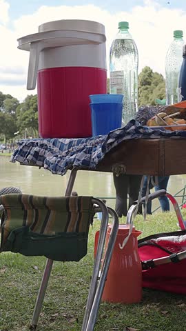 Some beverages on table in park for picnic in sunny day a small foldable chair and an orange jug rest on the grass, while a red bag is placed nearby. 